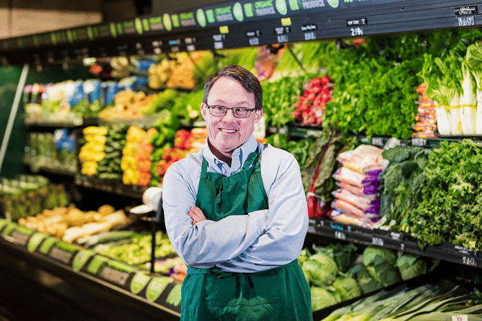 A man wearing a green apron standing with his arms cross, smiling in front of fruit and vegetables at a supermarket.