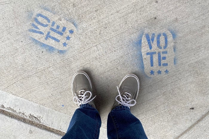 An aerial image of a person standing on concrete, with two blue graffiti artworks that read 'VOTE' and three stars beneath.
