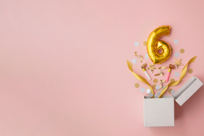 A gold number six balloon, popping out of a box, on a pink background.