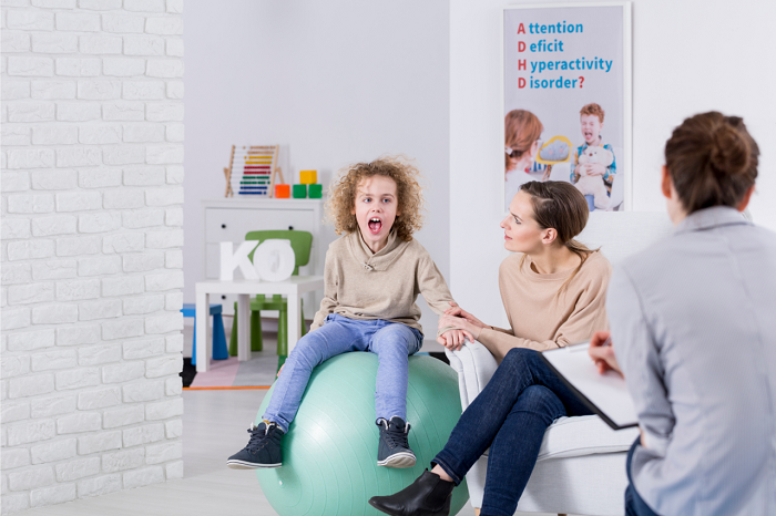 A child sitting on a exercise ball next to two adults in a classroom.