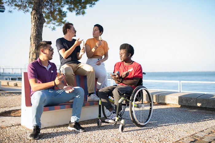 Four friends talking and eating ice cream on an outdoor bench. The ocean can be seen in the background.