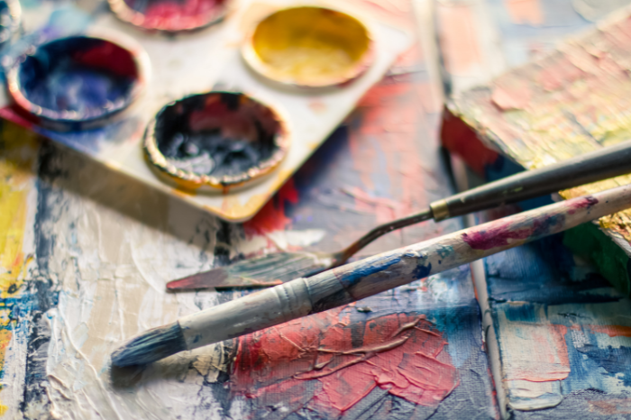 An artist's well-used paint brush, spatula and palette rest on top of a colourful canvas.