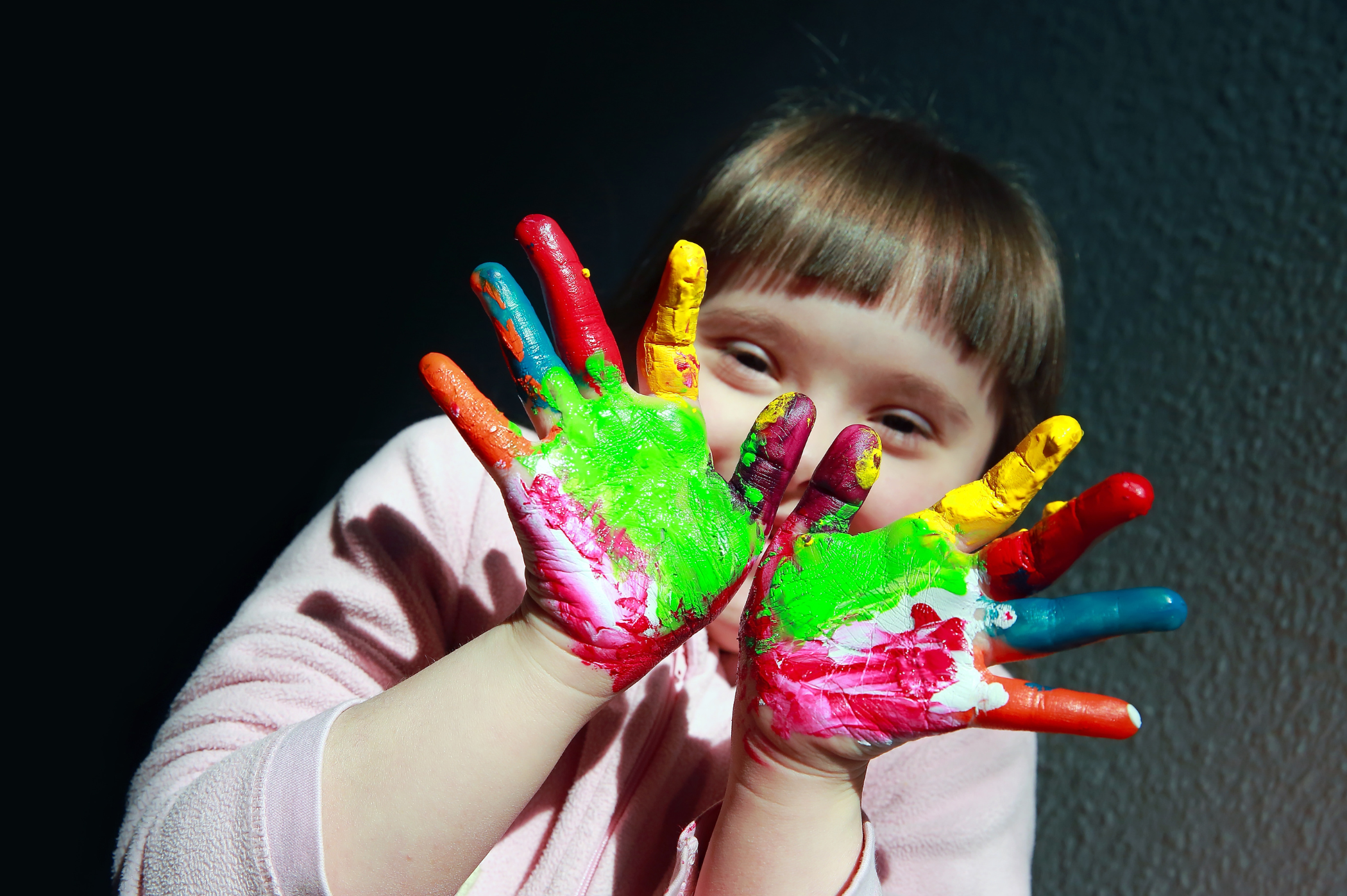 A little girl holds her hands up in front of her face. They're covered in brightly coloured paints.
