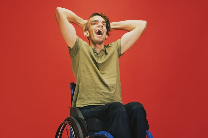 A young man with a trendy haircut and big smile sits in his wheelchair. The background behind him is red.