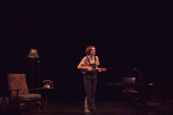 Creative producer and performer, Grace Colsey, performs with a ukulele on a dimly lit stage.